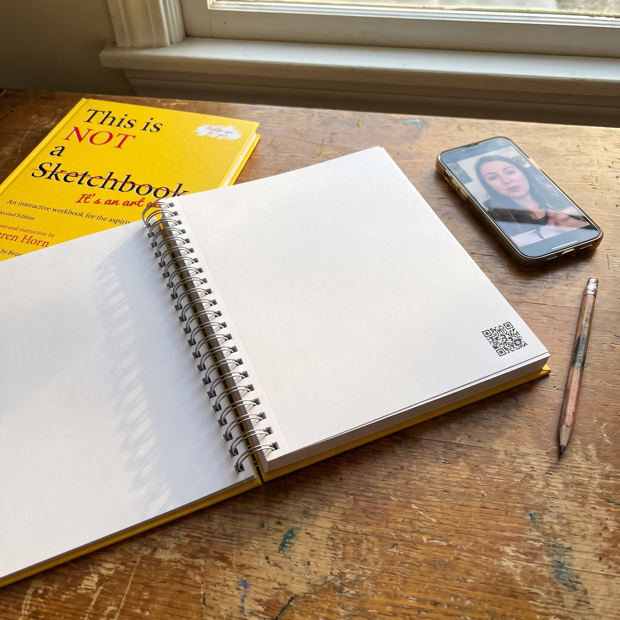 Open workbook on a wooden table beside a phone showing a paused video of an art teacher, with a pencil in frame.